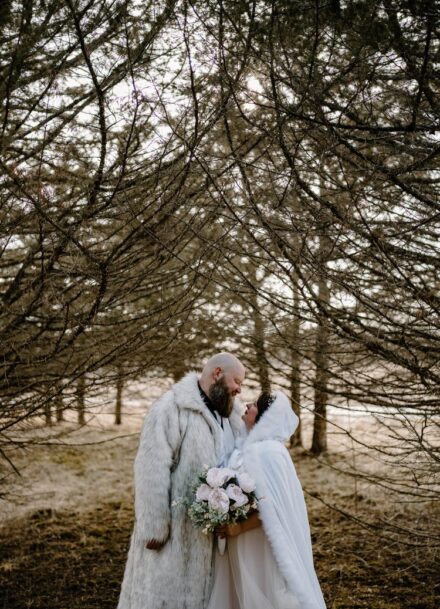 Lexi and Matt stand with their faces together underneath the branches of pine trees.