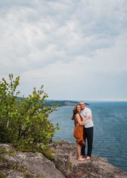 A couple stands on the edge of a cliff at Palisade Head, hair blowing in the wind.