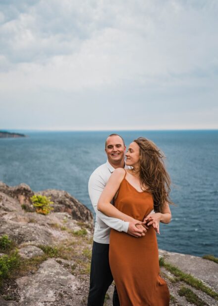 A couple stands on the cliff overlooking Palisade head, one of them being hugged from behind.