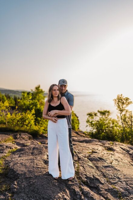 A couple stands with their arms wrapped around each other and Lake Superior in the distance.