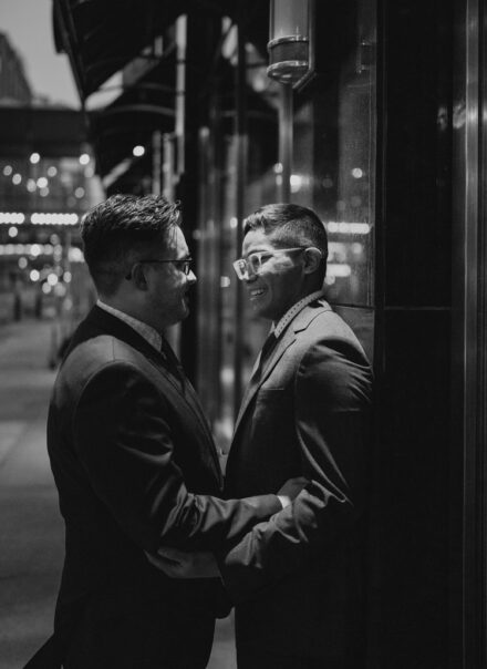 The grooms lean against the black marble facade of the restaurant, smiling at each other.