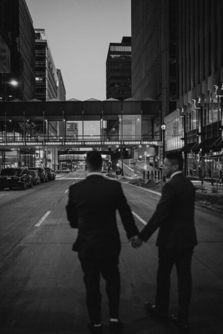 The grooms hold hands and walk into a city street. They are out of focus, but the buildings are clear.