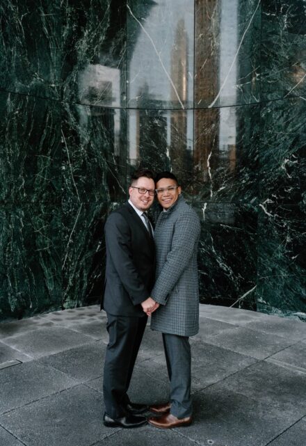 Zane and David hold hands and press the sides of their faces together in front of a green marble wall.