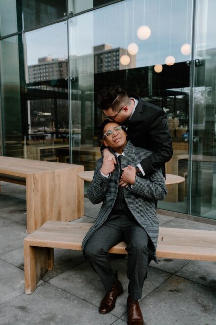 David sits on a wooden bench as Zane stands behind him, leaning him over to kiss David on the head.