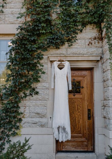 A Lulu's lace wedding dress hangs on an outdoor door frame on a brick wall covered in ivy.