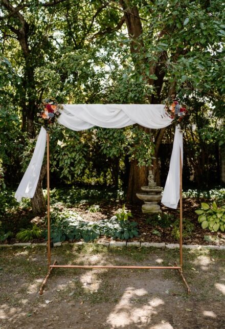 The copper wedding arch, draped in white fabric with wooden flowers in the top corners.