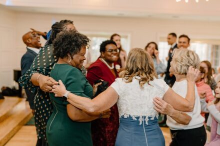 A group of wedding guests dances with their arms around each other.