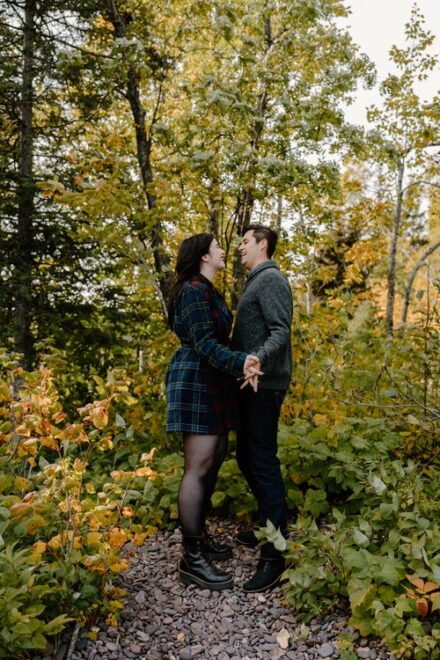 Alison and Sebastein hold hands facing each other in front of autumn leaf-covered trees.