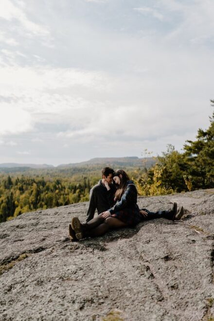 Alison and Sebastein sit with their legs facing opposite directions at an overlook, with autumn leaves in the distance.