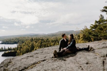 Alison and Sebastein sit with their legs facing opposite directions at an overlook, Alison's hand on Sebastein's face.