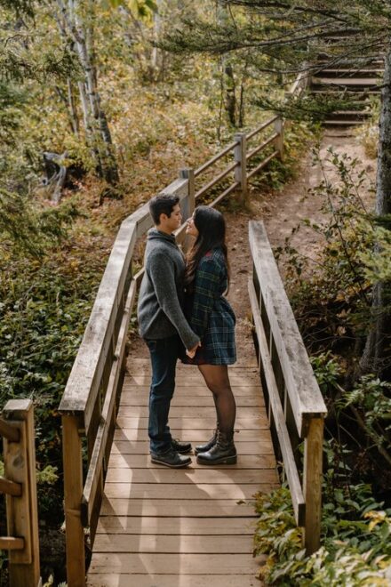 Alison and Sebastein stand in the middle of a wooden bridge surrounded by trees.