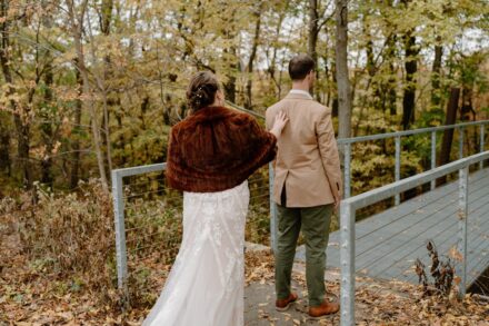 Laura taps on Joe's shoulder as he faces away from her on a bridge facing the forest.