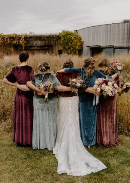 Laura and her bridesmaids in mismatched velvet dresses face away from the camera and wrap their arms around each other's shoulders.