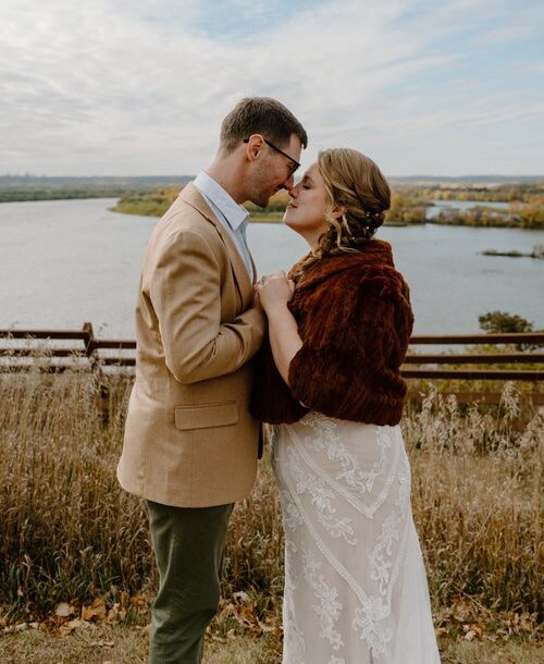 Laura and Joe hold hands and press their noses together in front of the view overlooking the river.