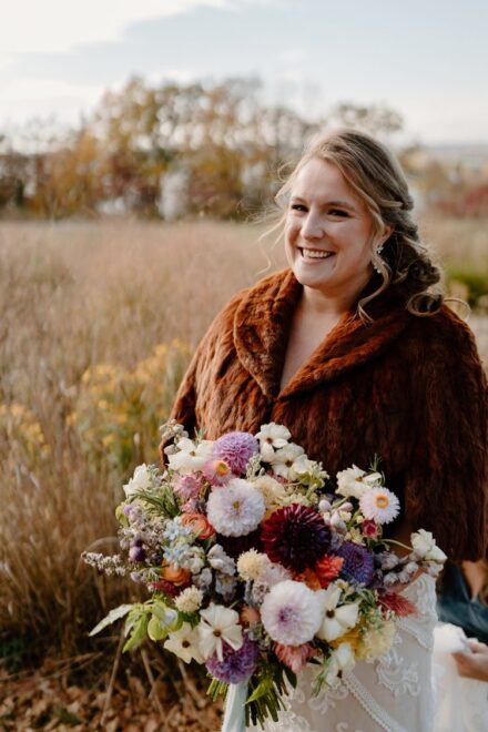 Laura holds her bouquet and smiles at the camera in front of a tall grass field.