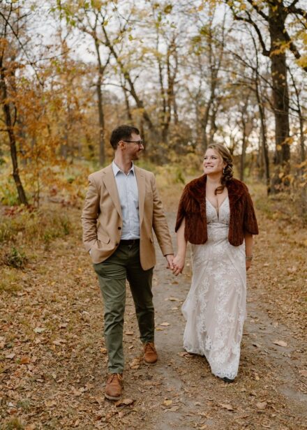 Laura and Joe walk down a dirt path surrounded by trees covered in fall leaves.