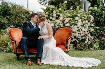 Sarah and Jake kiss sitting on an antique orange loveseat in front of a hydrangea bush.