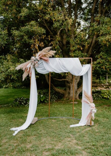 A copper wedding arch covered in white cloth and dried pink flowers.