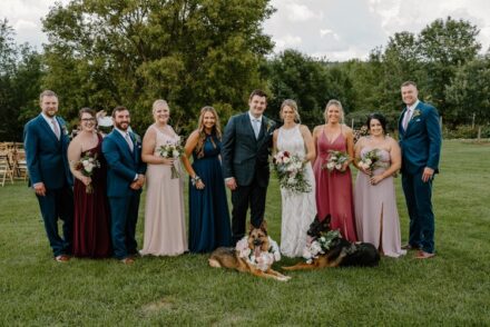 The wedding party casually stands in a grass field, smiling and staring at the camera with the two dogs sitting at their feet.