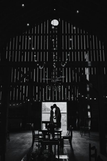 The bride and groom kiss in a doorway of a barn wall, with light shining through the slats of the wall.