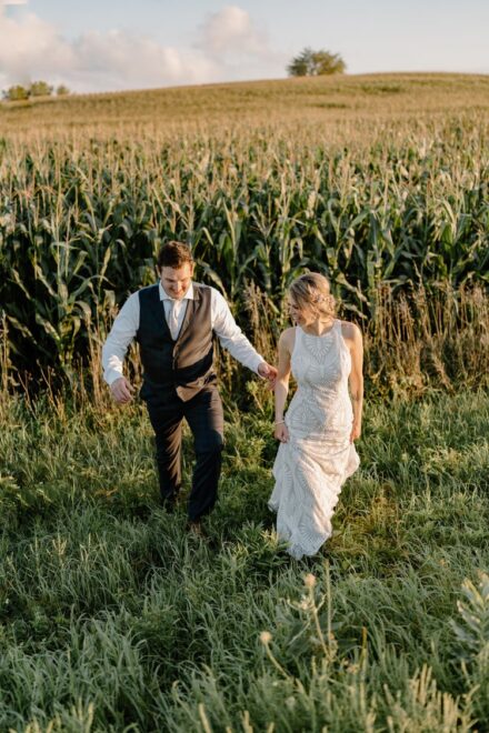The bride and groom walking together in the tall grass in front of a corn field during sunset.