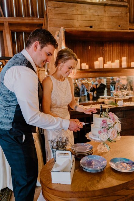 Sarah and Jake cut their small wedding cake with pink flowers under the golden lights of the barn.
