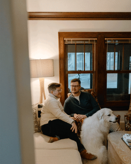 A gay couple sitting on a couch in their vintage styled living room with a Great Pyrenees at their feet.