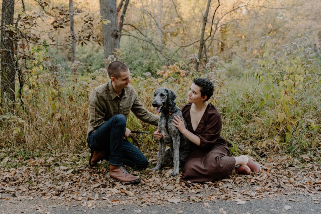 A couple sitting in a pile of fall leaves, looking at each other with their grey poodle sitting between them.