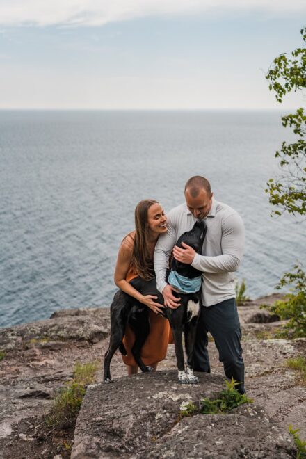 A couple standing on the cliffs of Palisade Head on Lake Superior hugging their black lab as she licks their faces.