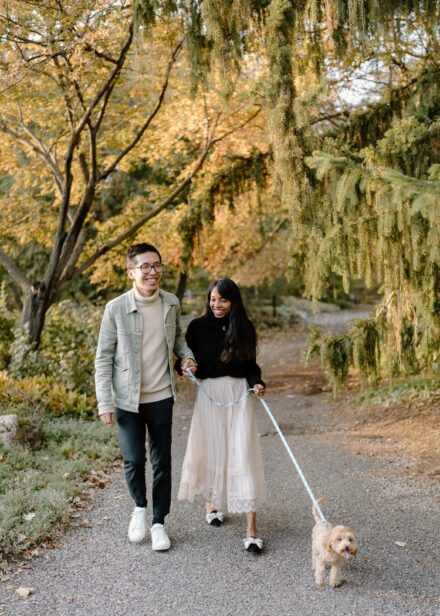 A couple walking their malti-poo puppy down a paved path surrounded by fall leaves and green, mossy trees.