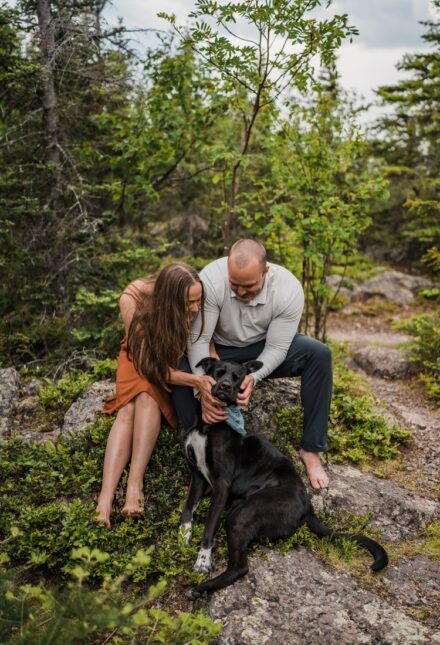 A couple sitting on a rock, smooshing the face of their happy black lab laying at their feet.