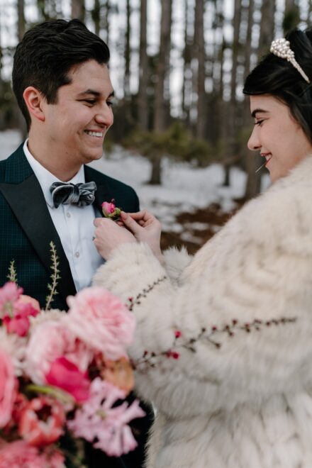 A bride in a white faux fur jacket pinning a boutonniere on the groom.