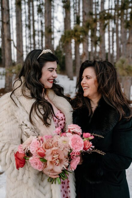 A bride in a pink wedding dress and white faux fur jacket smiling at her mom.