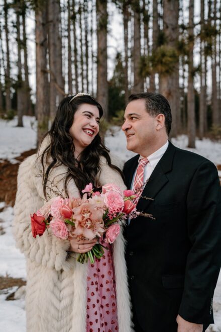 A bride in a pink wedding dress and white faux fur jacket smiling at her dad.