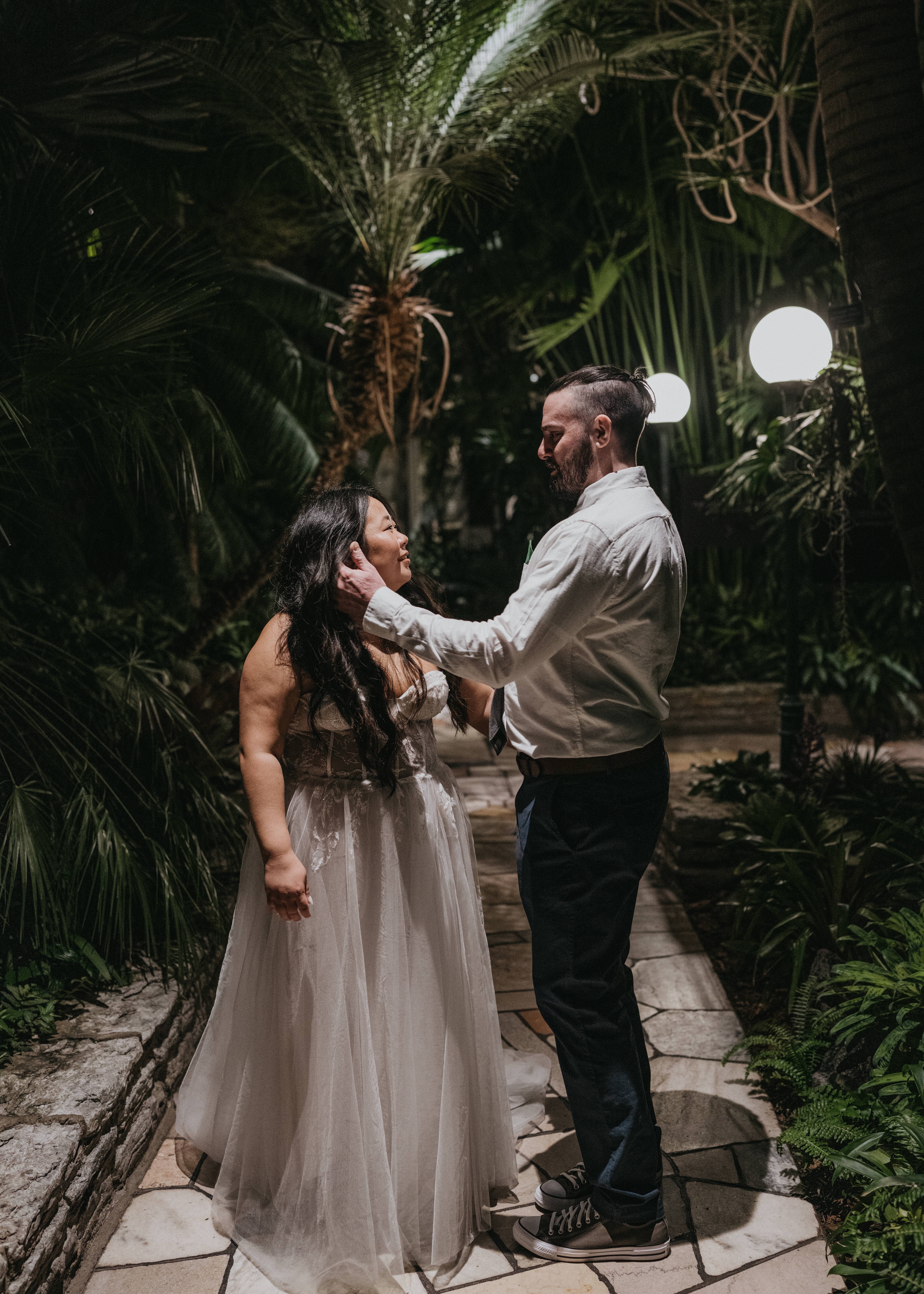 A wedding couple standing underneath palm trees at the Como Conservatory, facing each other and touching each other's faces.