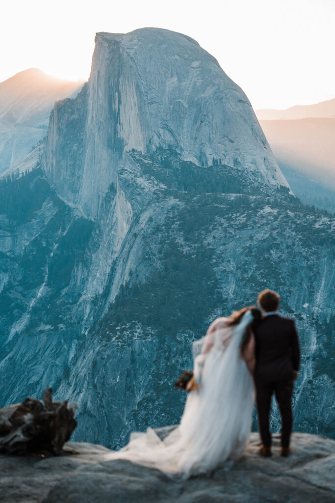 An elopement in Yosemite National Park. The couple in wedding attire is standing at Glacier Point, looking at Half Dome.