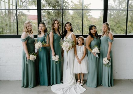 The bride and her bridesmaids in sage green wedding dresses, holding white floral bouquets.