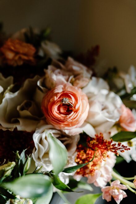 A diamond wedding ring tucked into the petals of a peach ranunculus in a flower bouquet.