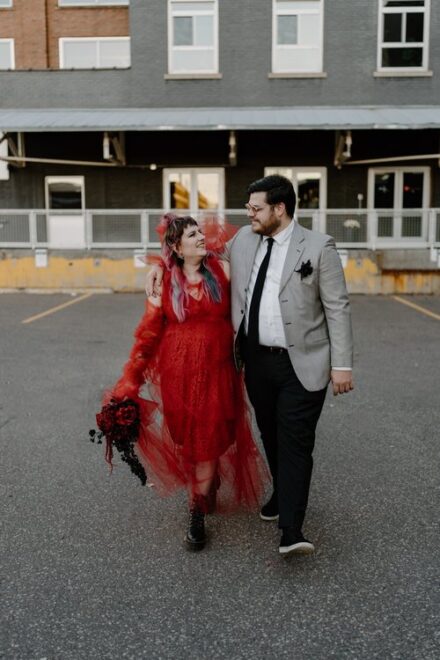 A couple in homemade Beetlejuice and Lydia Deetz costumes smile at each other as they walk toward the camera in a parking lot.
