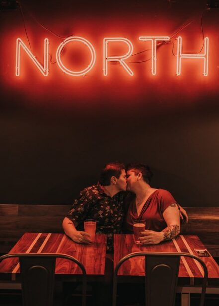 A lesbian couple kisses underneath a red neon sign that says NORTH.
