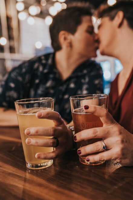Abby and Jill hold their beers with their left hands, showing off their engagement rings as they kiss out of focus in the background.