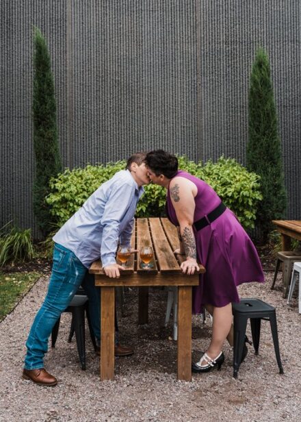 Abby and Jill stand on either side of a wooden table, leaning in for a kiss.