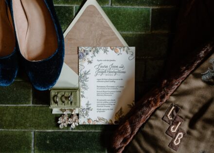 A flat lay of wedding details on jade green tile, including blue velvet heels, a velvet green ring box, and a vintage fur jacket.