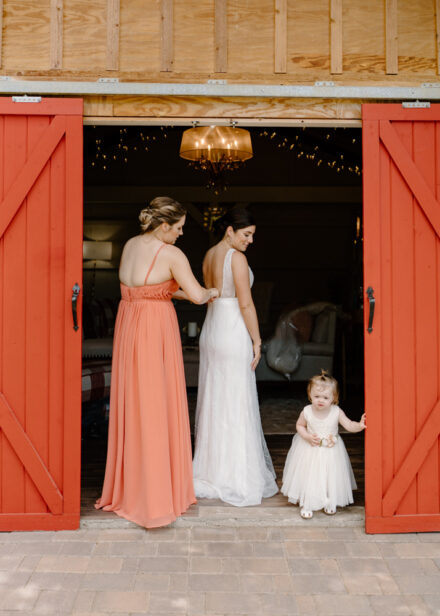 The maid of honor buttons up the back of Anna's dress between the red barn doors of the cabin.