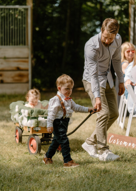 A friend guides the ring bearer down the aisle, helping him pull a wagon carrying the smallest flower girl.