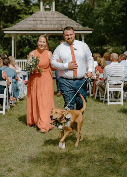 A bridesmaid and groomsman smile at the camera as they walk a dog down the aisle.