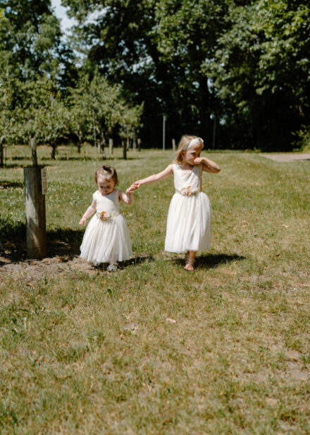 The two flower girls hold hands as they walk through the orchard.