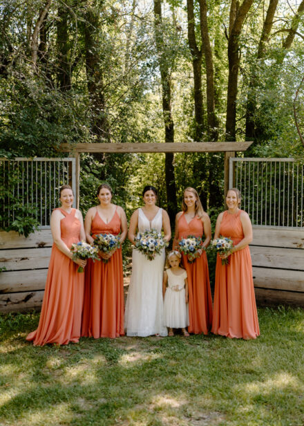 Bridesmaids in salmon pink dresses pose with the bride, holding white floral bouquets.