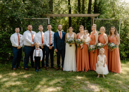The entire wedding party smiles sweetly for a camera, everyone in a line.
