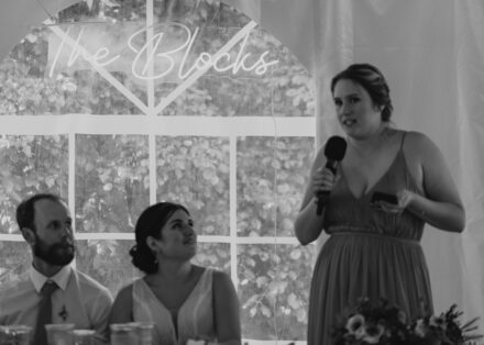 Anna and Jason smile at the maid of honor during her speech, a neon sign reading "The Blocks" hung above them.